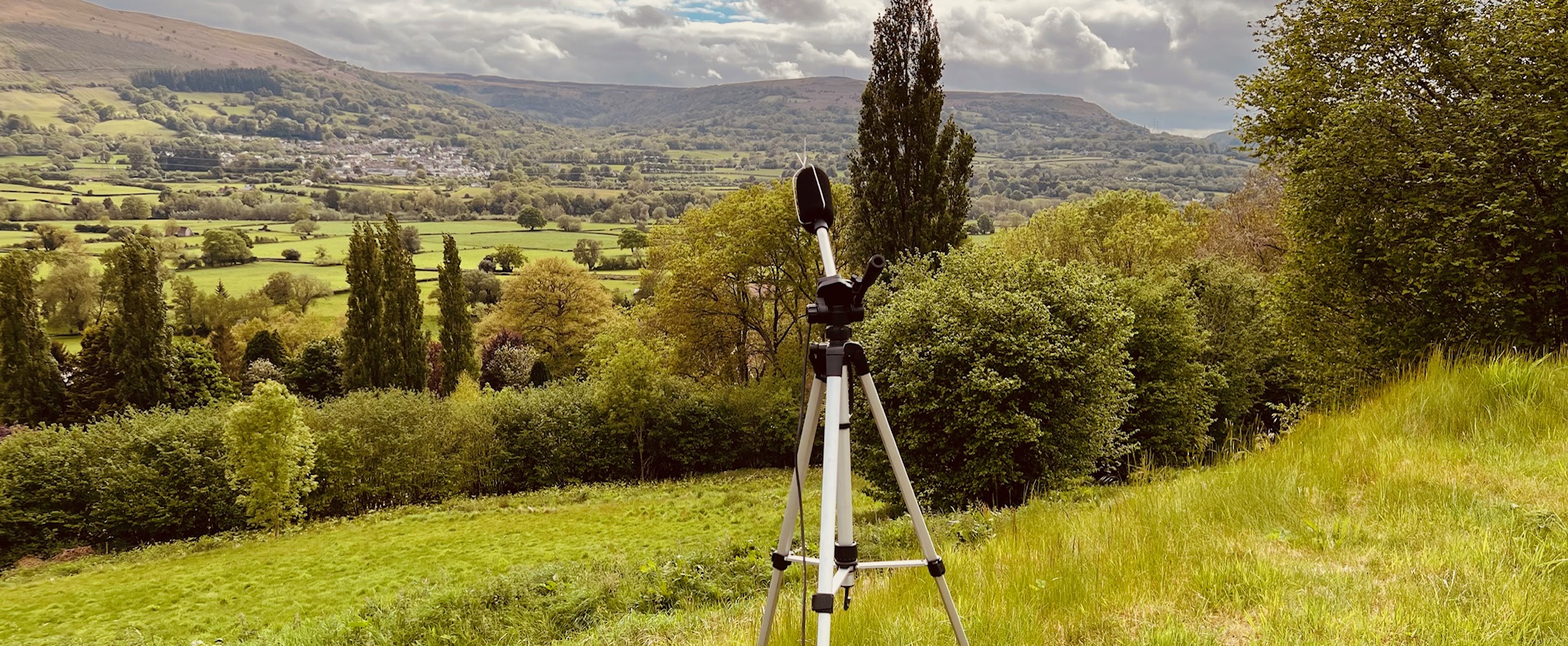 A tripod-mounted microphone setup in a scenic countryside, used for a noise survey as part of a noise impact assessment. This equipment helps gather data for BS4142 and BS8233 assessments to develop an effective noise management plan. ACP Acoustic Consultants